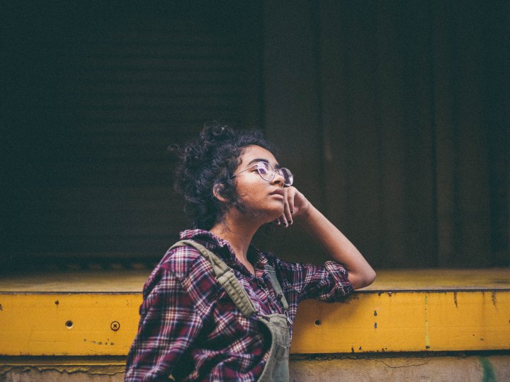 A young woman of color wearing glasses and a flannel shirt leans against a cement structure while looking wistfully to the right.