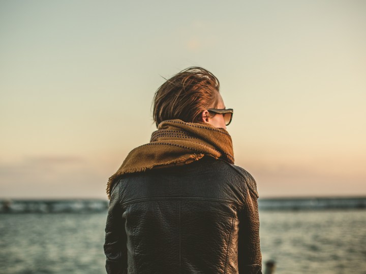A person wearing a leather jacket, a scarf, and sunglasses is looking out over the ocean on a cloudy day.