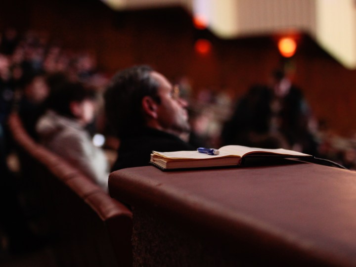 An audience sitting in some kind of theather are in the background. An open book with a pen sitting on top of it is in the foreground.